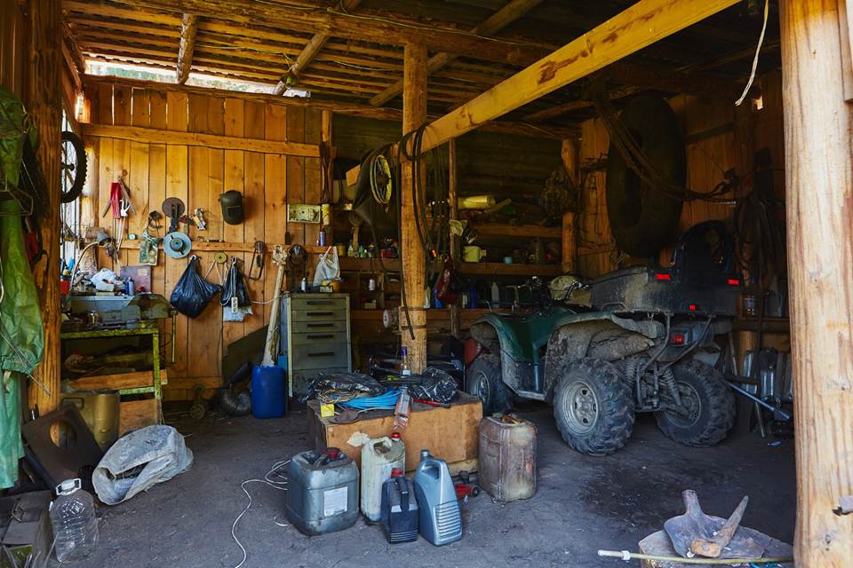 A cluttered wooden garage with tools, jugs, bags, and spare parts scattered and hanging on walls. An ATV is parked inside, surrounded by equipment and supplies in a rustic, dimly lit space.